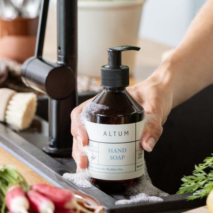 Hånd holder ALTUM Golden Grass håndsæbe 250 ml fra Ib Laursen ved køkkenvask