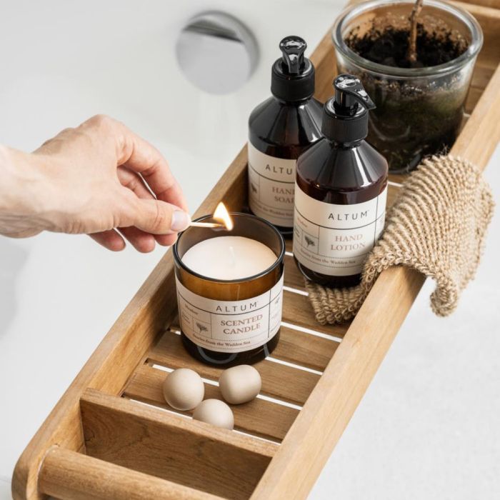 En hånd tænder et Ib Laursen ALTUM Meadow duftlys i brunt glas på bakke over badekar