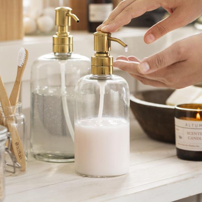 En hånd trykker på Ib Laursen sæbedispenser glas og messingfarvet pumpe med hvid sæbe