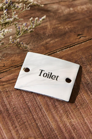 Hvidt skilt i emalje med teksten Toilet fra Ib Laursen liggende på gulv