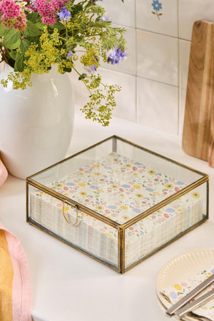 Solitaire glasæske med guldfarvet ramme og servietter på et lyst bord ved en vase med blomster